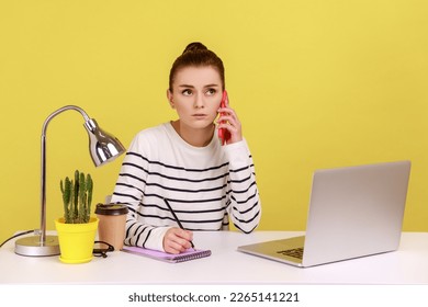 Concentrated secretary in striped shirt receiving calls on phone and making notes, writing down information, sitting at workplace with laptop. Indoor studio studio shot isolated on yellow background. - Powered by Shutterstock