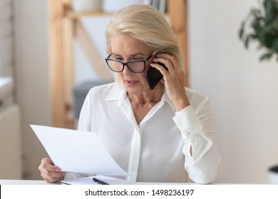 Concentrated senior businesswoman in glasses sit at office desk talk on cellphone reading paperwork document, focused middle-aged woman speak with client on smartphone discuss contract - Powered by Shutterstock