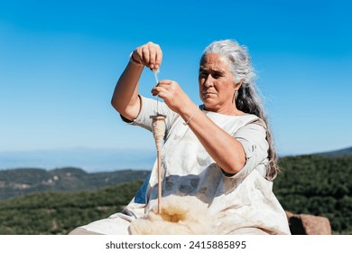 Concentrated senior female sitting under blue sky and making thread with thread maker spinning wheel while working against picturesque mountains view - Powered by Shutterstock