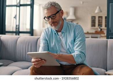 Concentrated senior man in casual clothing using digital tablet while sitting on the sofa at home - Powered by Shutterstock