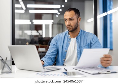 Concentrated and serious Arab young man sitting in the office and working with data, typing on a laptop and holding documents in his hands. - Powered by Shutterstock