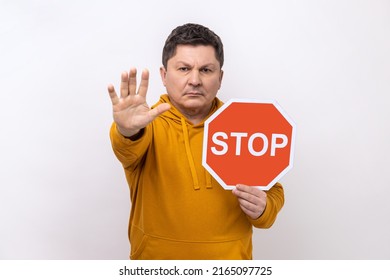 Concentrated serious man looking angrily showing stop gesture, holding road traffic sign, warning of ban, forbidden access, wearing urban style hoodie. Indoor studio shot isolated on white background.