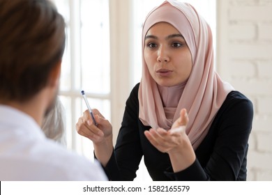 Concentrated serious young arabian businesswoman in hijab talking to male team leader, headshot. Focused mixed race partners discussing marketing strategy or project details together at office. - Powered by Shutterstock