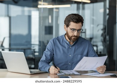 Concentrated and serious young male accountant sitting in the office behind a desk and working with documents, checking and analyzing information and data. - Powered by Shutterstock