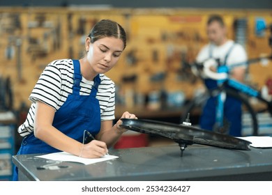 Concentrated skilled young female mechanic attentively examining bike wheel rim and documenting service checklist at counter of bicycle repair workshop - Powered by Shutterstock