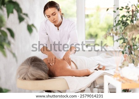 Concentrated skilled young masseuse doing back pain relief massage to elderly female client lying on massage table in medical office..