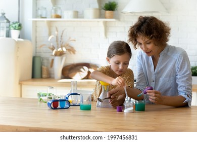 Concentrated small kid girl playing with young mom, involved in doing chemistry experiments together in modern kitchen. Happy mother educating little child daughter at home, making biological tests.