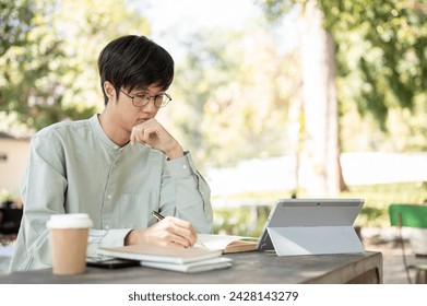 A concentrated and smart Asian male university student is doing homework or studying online on his digital tablet while sitting at a table in a park. - Powered by Shutterstock