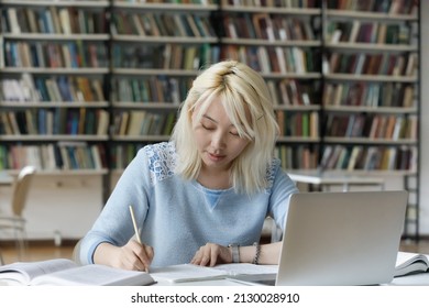 Concentrated Smart Pretty Asian Female Student Writing Notes In Copybook, Reading Books And Watching Educational Online Lecture On Computer, Sitting At Table Alone In Modern College Library.