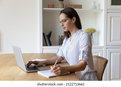Concentrated smart young woman in eyeglasses web surfing useful information, improving knowledge studying on online courses, writing notes in copybook, preparing for examination at home office. - Shutterstock ID 2122713662