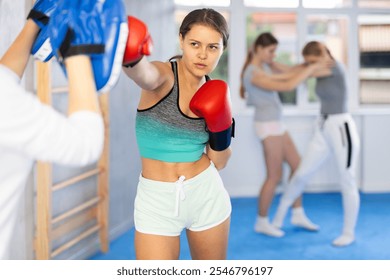 Concentrated sporty teen girl in boxing gloves practicing self-defense techniques in gym, throwing punches on focus mitts in hands of instructor.. - Powered by Shutterstock