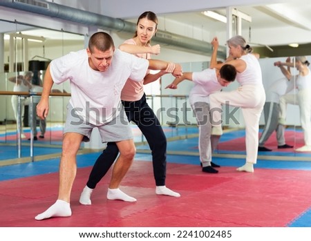 Concentrated sporty young woman learning self defence techniques in sparring with man, practicing elbow blow with wristlock to opponent in gym