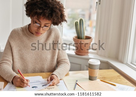 Concentrated statistician studies statistics, reads information data, examines chart, drinks coffee from disposable cup. Busy dark skinned architect works from home. People and documentation concept