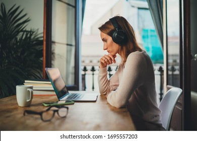 Concentrated student girl learning online having video call via laptop computer sitting at home interior near open balcony, female entrepreneur working remotely from home office using modern computer