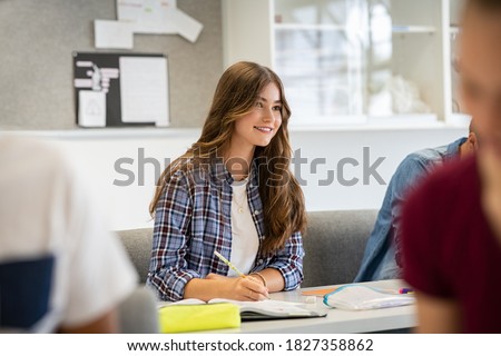 Concentrated student studying and taking notes in a notebook sitting in lecture during lesson. Smiling high school teen sitting in classroom with other students listening to professor. Happy girl.