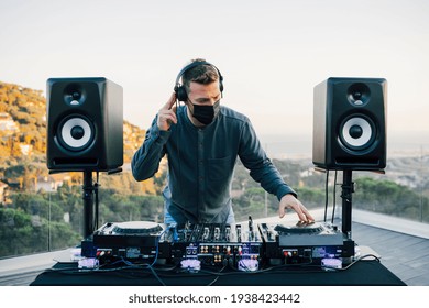 Concentrated Techno Young Caucasian Male DJ In Surgical Mask And Headphones Playing Music At A Home Terrace During Sunset, Mixing At His Mixer Board, During Coronavirus Pandemic, Quarantine Party.