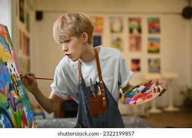 Concentrated teenage asian gay man dressed in apron painting picture with oil paints on canvas in bright art class