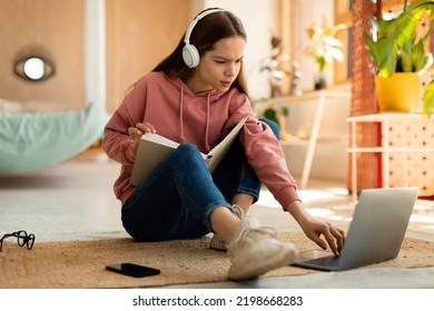 Concentrated teenage girl reading book in front of laptop, getting ready for exam, sitting on floor at home. Focused female student having remote lesson on pc