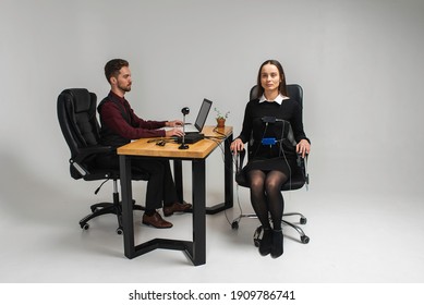 Concentrated, Thoughtful Woman Is In A Bright Room, Testing On A Computer Polygraph. Young Man Sitting At The Table And Looking At The Polygraph Screen And Polygraph Monitoring. The Concept Of Truth, 