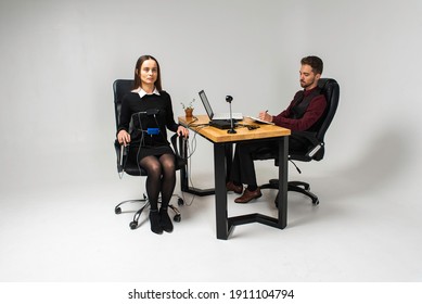 Concentrated, Thoughtful Woman Is In A Bright Room, Testing On A Computer Polygraph. Young Man Sitting At The Table And Looking At The Polygraph Screen And Polygraph Monitoring. The Concept Of Truth