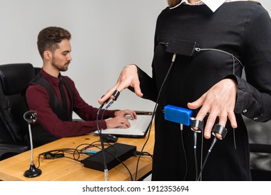 Concentrated, Thoughtful Woman Is In A Bright Room, Testing On A Computer Polygraph. Young Man Sitting At The Table And Looking At The Polygraph Screen And Polygraph Monitoring. The Concept Of Truth