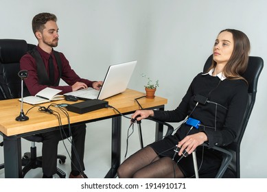 Concentrated, Thoughtful Woman Is In A Bright Room, Testing On A Computer Polygraph. Young Man Sitting At The Table And Looking At The Polygraph Screen And Polygraph Monitoring. The Concept Of Truth