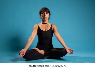Concentrated trainer meditating during pilates workout sitting in lotus position on yoga mat practicing spiritual meditation. Gymnast woman training body muscles in studio with blue background