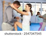 Concentrated tween girl engaging training sparring with instructor during group self-defense course, performing knee strike in midsection to man ..