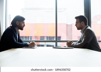 Concentrated two businessmen sitting at table, involved in serious negotiations or contract discussion in modern office. Focused young male ceo executive manager communicating with partner or client.