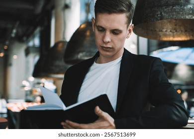 Concentrated well dressed male in black suit sitting near table and reading book while resting in modern cafeteria during break