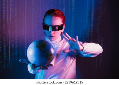 Concentrated woman in futuristic holographic clothes and VR glasses holding earth globe model on the one hand and affecting on it by another hand with matrix code background. Neon blue and pink light.