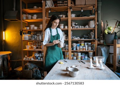 Concentrated woman potter connecting handle to clay mug for secure material bonding, preparing ceramic piece for next stage of drying and firing in pottery workshop. Ceramist at work in store studio - Powered by Shutterstock