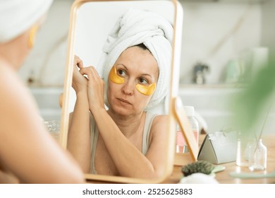 Concentrated woman with towel tied around head using hydrating collagen under-eye patches, preparing for day in front of mirror - Powered by Shutterstock