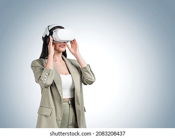 Concentrated Woman With Virtual Reality Glasses Standing In Beige Suit. Concept Of Future And Imaging Technology. Copy Space Empty Blue Wall