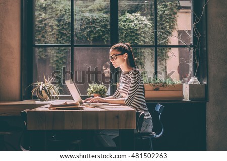 Concentrated at work. Confident young woman in smart casual wear working on laptop while sitting near window in creative office or cafe