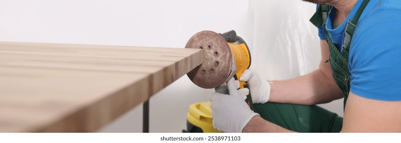 Concentrated worker polishes wooden board edge with grinding tool in room. Handyman processes timber detail in carpentry workshop. Furniture production - Powered by Shutterstock