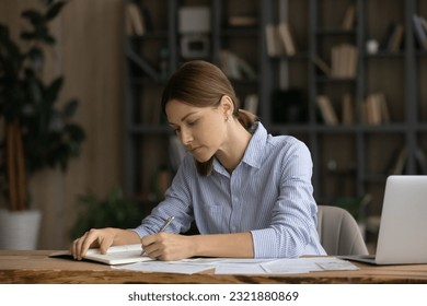 Concentrated young 30s caucasian woman writing business notes in paper organizer, planning future investments or managing monthly budget, reviewing bills or taxes, sitting at table at home office. - Powered by Shutterstock