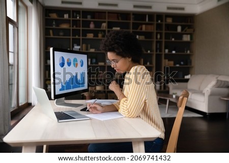 Concentrated young African American businesswoman employee worker in eyeglasses analyzing online sales statistics data on computer, reviewing marketing research, working with documents at home office.