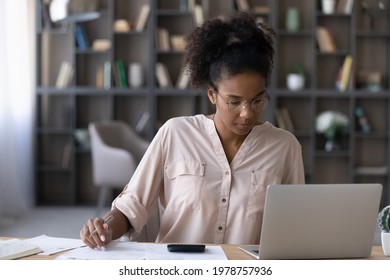 Concentrated young african ethnicity woman in eyeglasses calculating domestic expenditures, managing monthly household budget, paying utility bills or taxes online using computer e-banking application - Powered by Shutterstock