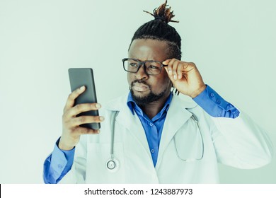 Concentrated young African male doctor reading message on phone and adjusting glasses. Serious hipster medical specialist with Afro hairstyle using gadget. Modern medicine concept - Powered by Shutterstock