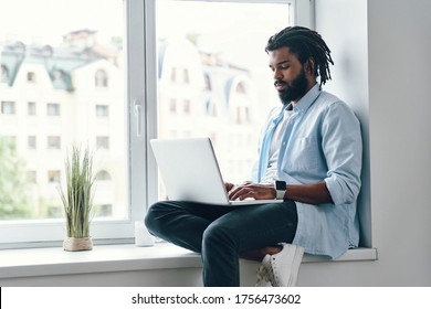 Concentrated young African man using laptop while sitting on the window sill indoors       - Shutterstock ID 1756473602