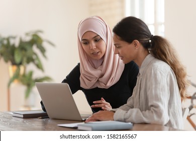 Concentrated young arabian woman in hijab sitting with smiling colleague at table, looking at computer screen, explaining new company software. Focused team leader training millennial female intern.