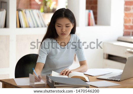 Concentrated young Asian female student sitting at desk in library. Focused millennial girl studying, preparing for entrance exams or university tests, reading book, taking down notes in textbook.