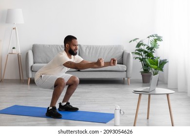 Concentrated young black bearded athlete man in white sports clothes squatting in living room interior, profile. Fitness blog, social distance, device, new normal and workout at home during covid-19