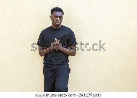 Concentrated young black male millennial in casual wear standing against pale yellow wall on street and looking at camera with attention
