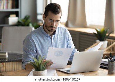 Concentrated young businessman ceo manager in eyewear looking at marketing research report, analyzing statistics data in charts, developing growth strategy, working on computer alone in modern office.