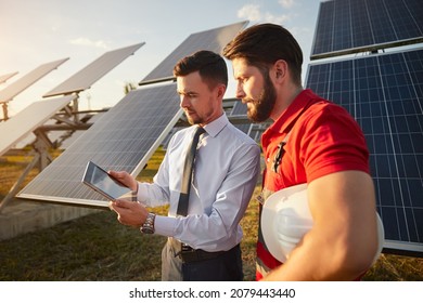 Concentrated Young Businessman In Formal Outfit Showing Project Details On Tablet To Male Technician Standing Near Photovoltaic Panels On Solar Power Station