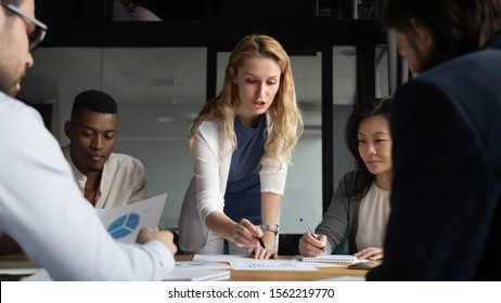 Concentrated young businesswoman explaining market research results in graphs to mixed race colleagues. Focused group of diverse employees holding brainstorming meeting, discussing project ideas.