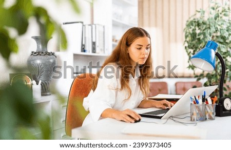 Concentrated young businesswoman working with laptop and papers at office desk..