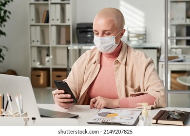 Concentrated Young Caucasian Bald Woman In Mask Sitting With Smartphone At Table While Working With Business Papers In Office